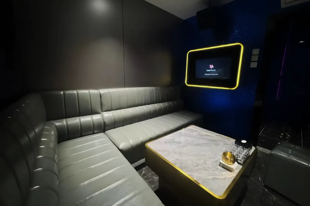 A sleek karaoke room features a curved gray leather sofa, a marble-topped table, and a wall-mounted screen with a yellow glow, set against a blue accent wall.