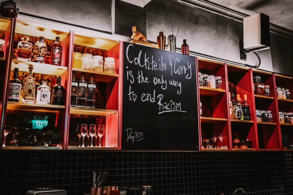 Bar shelves with various liquor bottles and glassware, featuring a blackboard with the message: "Cocktails & Unity is the only way to end Racism."