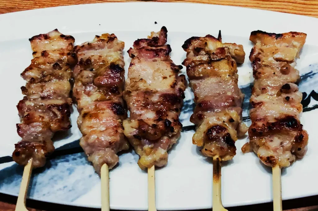 Five grilled skewers of seasoned chicken are neatly arranged on a white plate. The meat appears juicy and lightly charred, evoking a savory aroma.