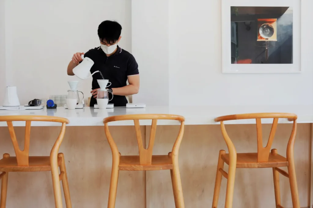 Barista in a mask makes pour-over coffee in a minimalist café. Three wooden chairs line the counter, and a modern art piece hangs on the white wall.
