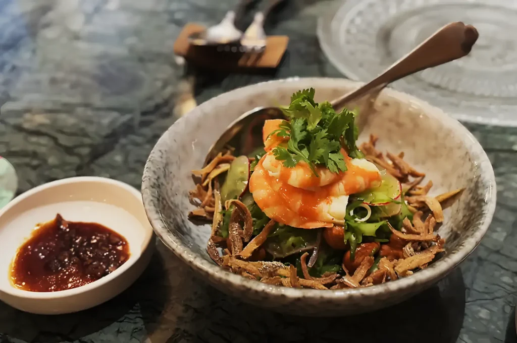 Bowl of shrimp salad with greens, garnished with cilantro and crispy toppings. A side dish of chili sauce is on the table. The setting is casual.
