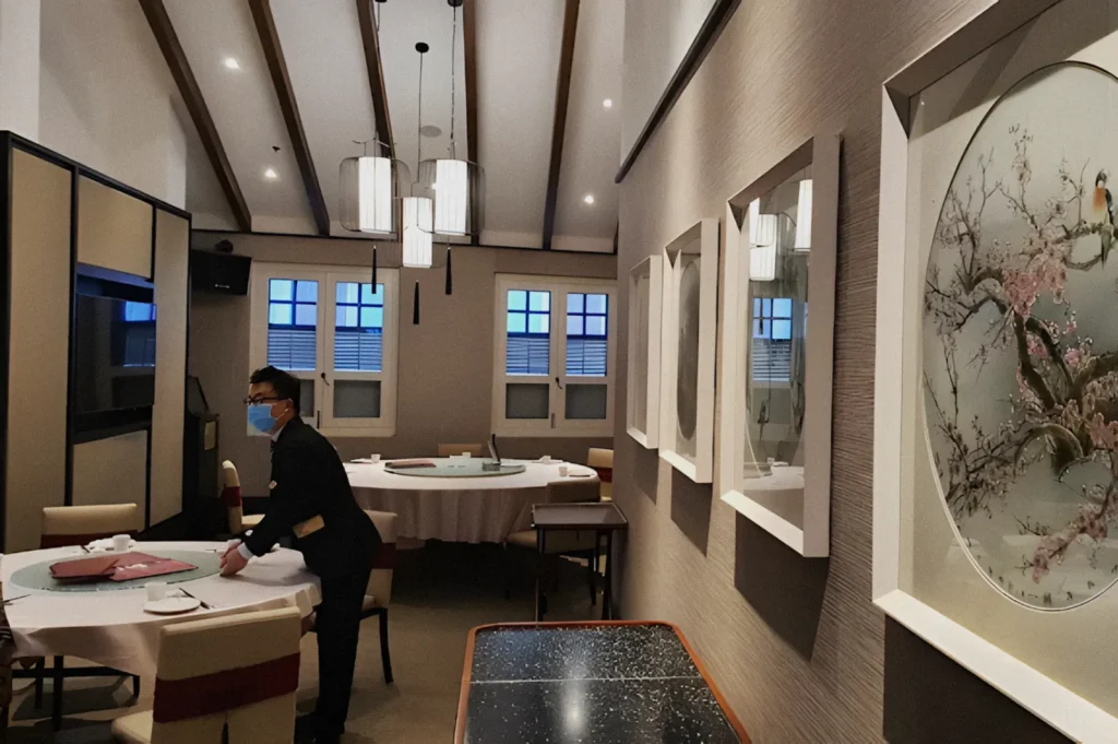 A waiter in a mask sets a table in an elegant restaurant with high ceilings and soft lighting. Wall art of birds and blossoms adds a serene touch.