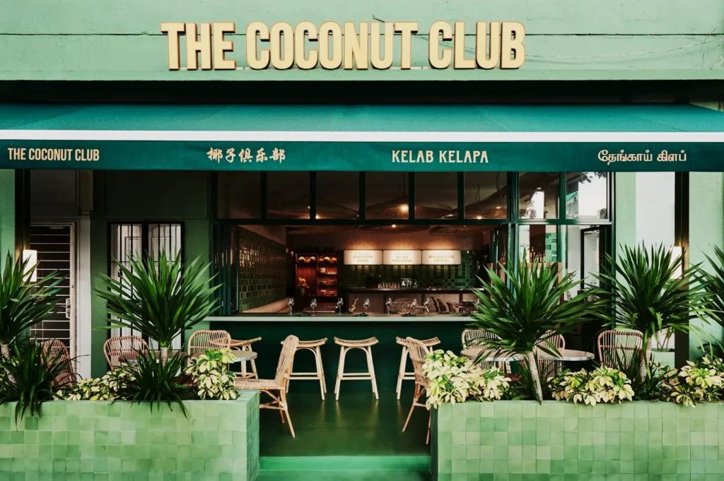 The street view of The Coconut Club café shows a vibrant green façade, with wicker chairs and tables on the patio. Lush plants add a tropical feel.