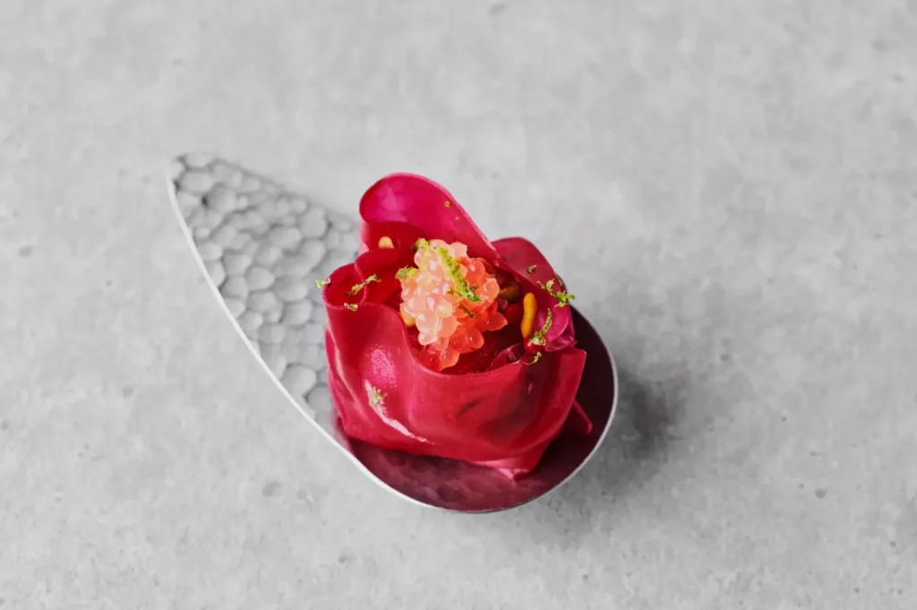 Elegant dish on a textured silver spoon, featuring vibrant pink petals filled with delicate, translucent pink beads, garnished with green zest.