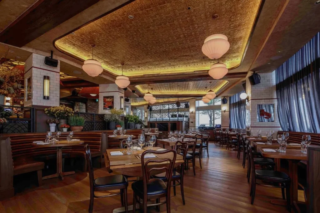 Warmly lit restaurant interior with wooden tables set for dining. Ornate ceiling, cozy booths, and blue curtains create a welcoming, elegant atmosphere.