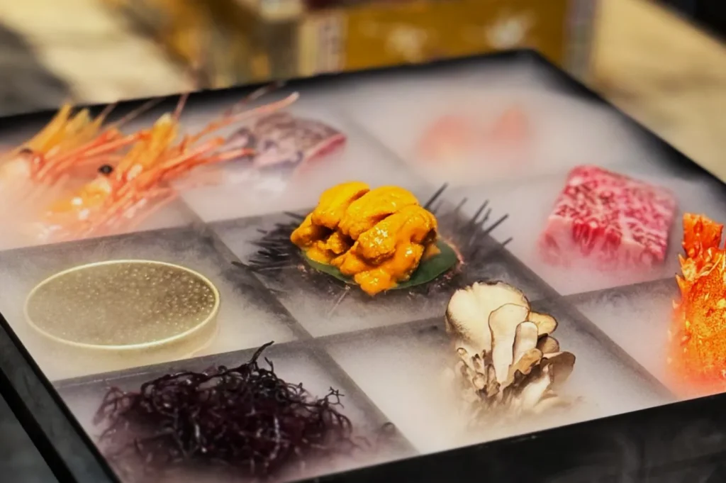 A nine-section tray features seafood and mushrooms artfully arranged. Dry ice smoke adds a dramatic flair, highlighting vibrant colors and textures.