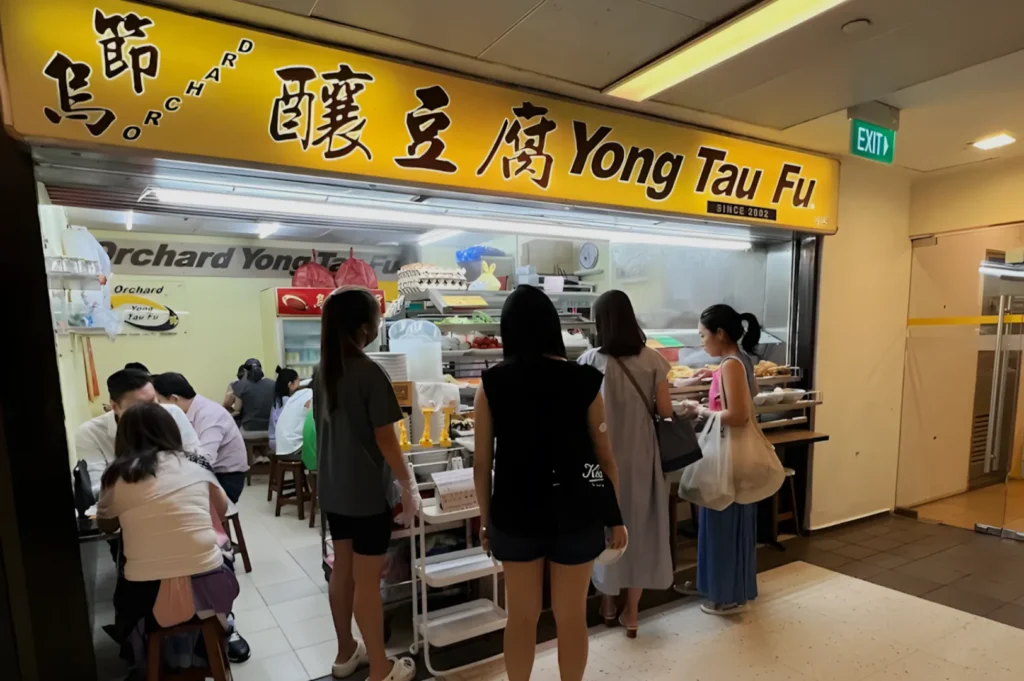 Customers line up at an Orchard Yong Tau Fu stall under a bright yellow sign. Inside, people eat at small tables, creating a lively and bustling atmosphere.