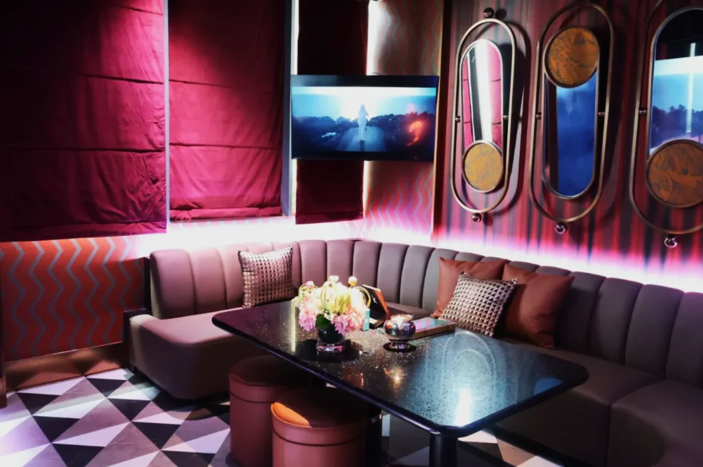 A cozy lounge with maroon drapes, a plush corner sofa, and geometric flooring. A TV shows a scene beneath soft lighting, with elegant pink flowers on the table.