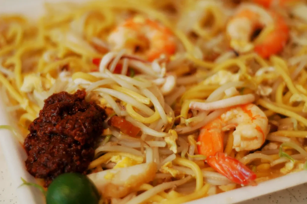 A dish of Hokkien Mee features yellow noodles, prawns, bean sprouts, and a dollop of sambal. A lime is visible on the side, adding a fresh touch.