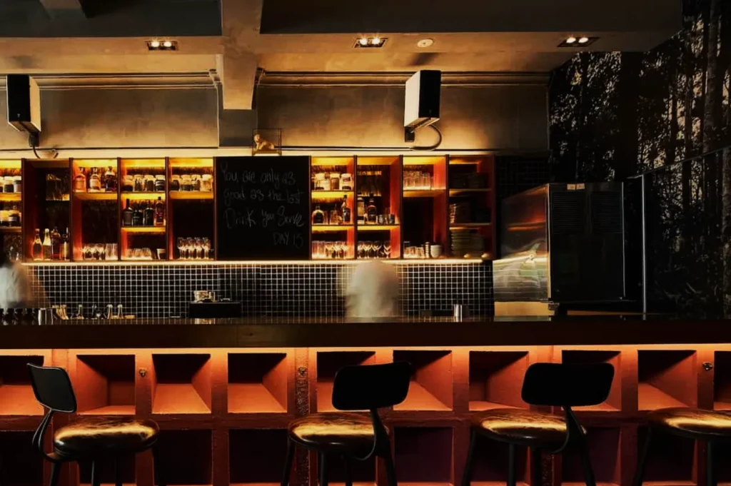 Cozy modern bar with warm lighting, four plush stools, a dark wood counter, and a stocked shelf of bottles. Chalkboard with a motivational quote adds character.