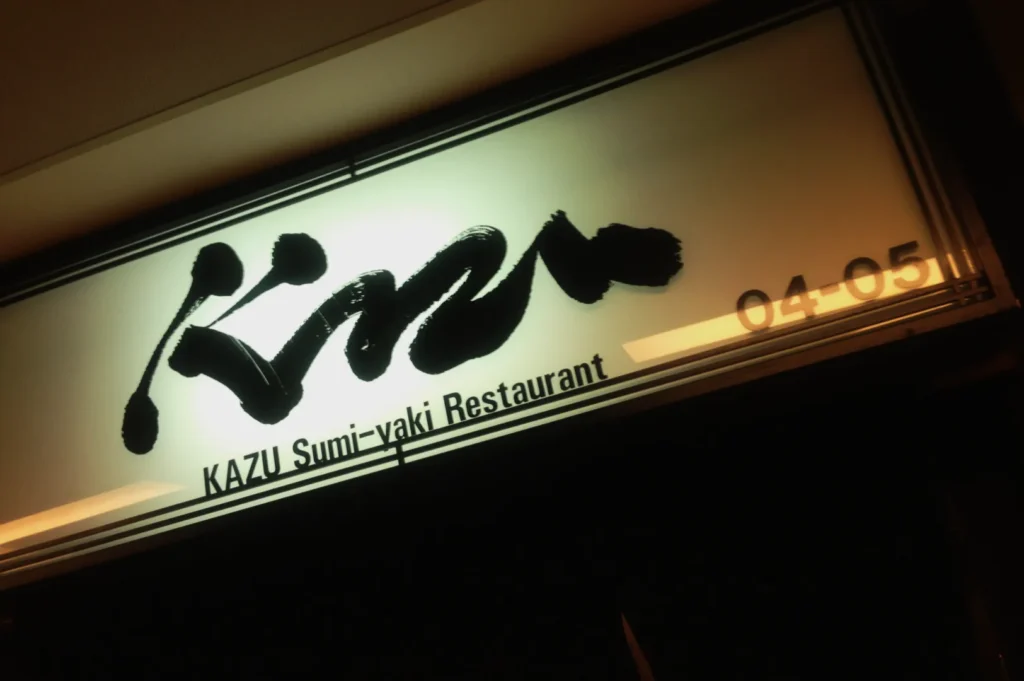 Illuminated restaurant sign reads "KAZU Sumi-yaki Restaurant" in black calligraphy, set against a light background. Warm ambiance conveyed.