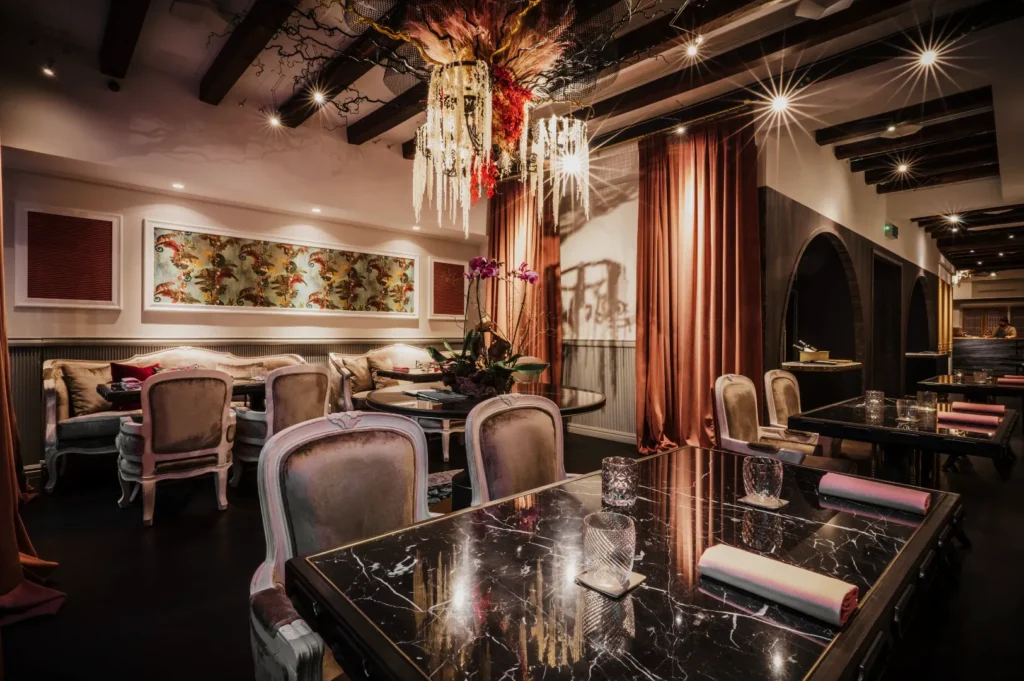 Elegant restaurant interior with marble tables, ornate chandeliers, vintage chairs, and warm lighting. Walls feature artistic panels and lush drapery, creating a cozy ambiance.