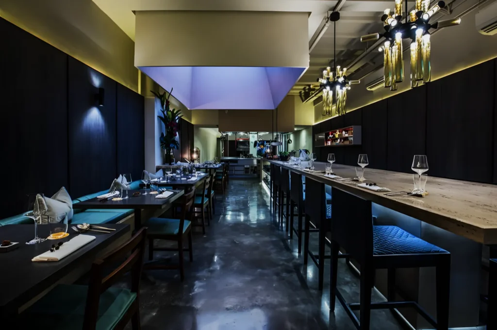 Sleek restaurant interior with dim lighting, dark wood walls, and elegant chandeliers. Tables set with glasses and napkins, creating an upscale ambiance.