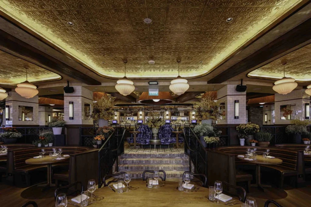 Elegant restaurant interior with dim lighting, featuring ornately designed ceilings, cozy booths, warm wood tones, plush seating, and decorative plants.