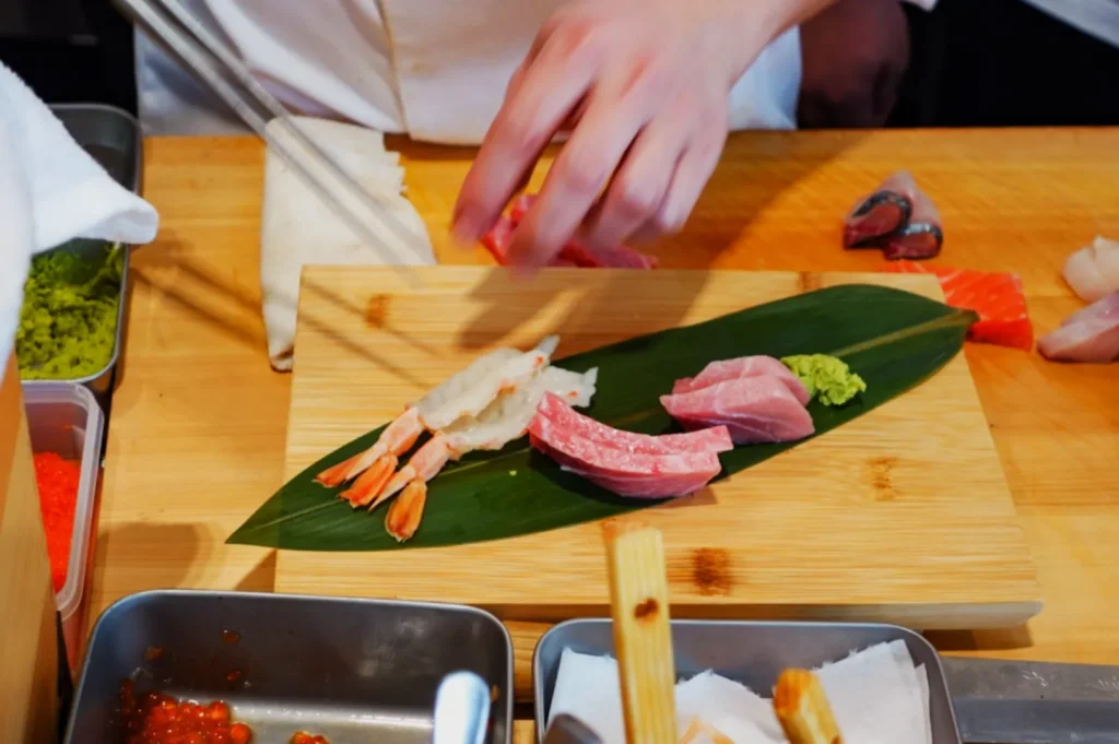 Chef's hands preparing sushi on a wooden board with a leaf. Includes shrimp, tuna, and wasabi, surrounded by vibrant ingredients. Elegant and focused.