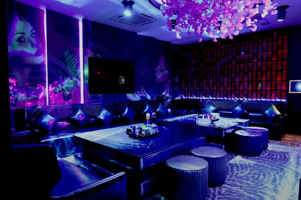 A vibrant karaoke lounge with purple and blue lighting, leather seating, and a table with microphones. Cherry blossom decorations hang overhead, creating a lively atmosphere.