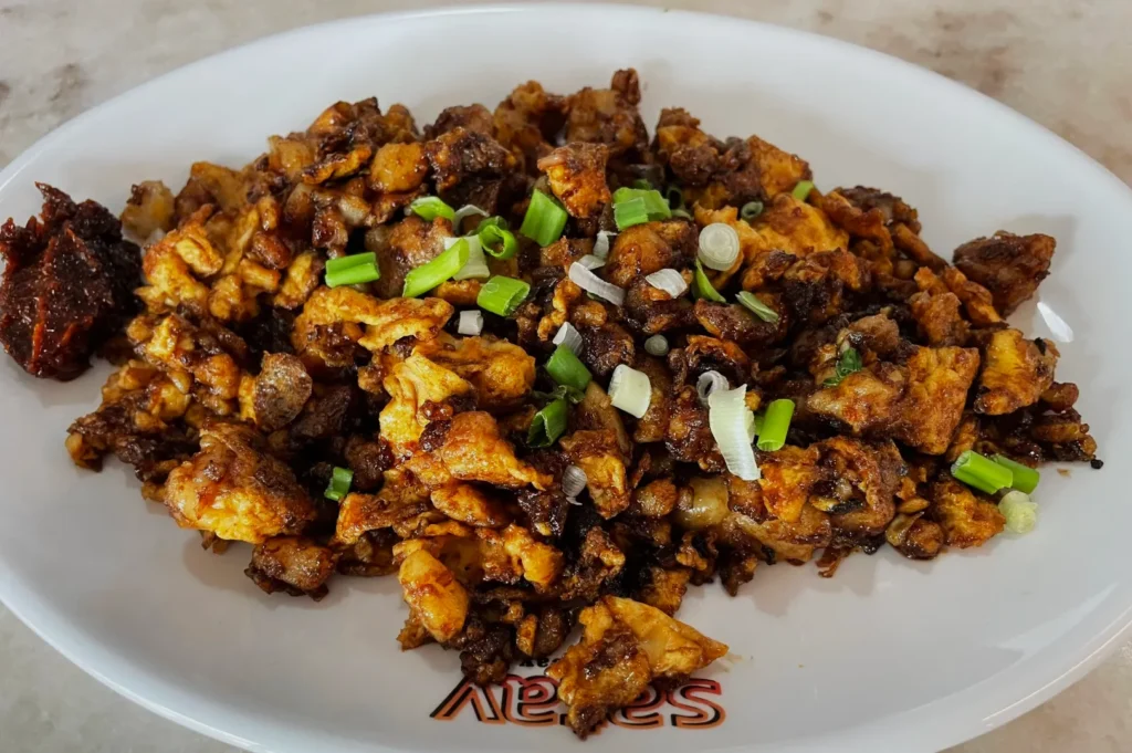 Stir-fried carrot cake dish on a white plate, garnished with green onions. It appears flavorful with a hint of spiciness, offering a savory aroma.