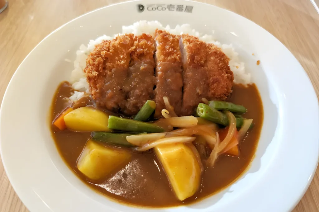 A plate of katsu curry features crispy breaded cutlet on white rice, topped with rich brown curry sauce, alongside vibrant vegetables.