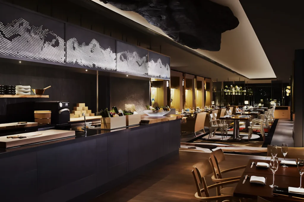 Sleek modern restaurant with wooden tables, elegant chairs, and ambient lighting. An open kitchen features a sculptural ceiling and art on the walls.