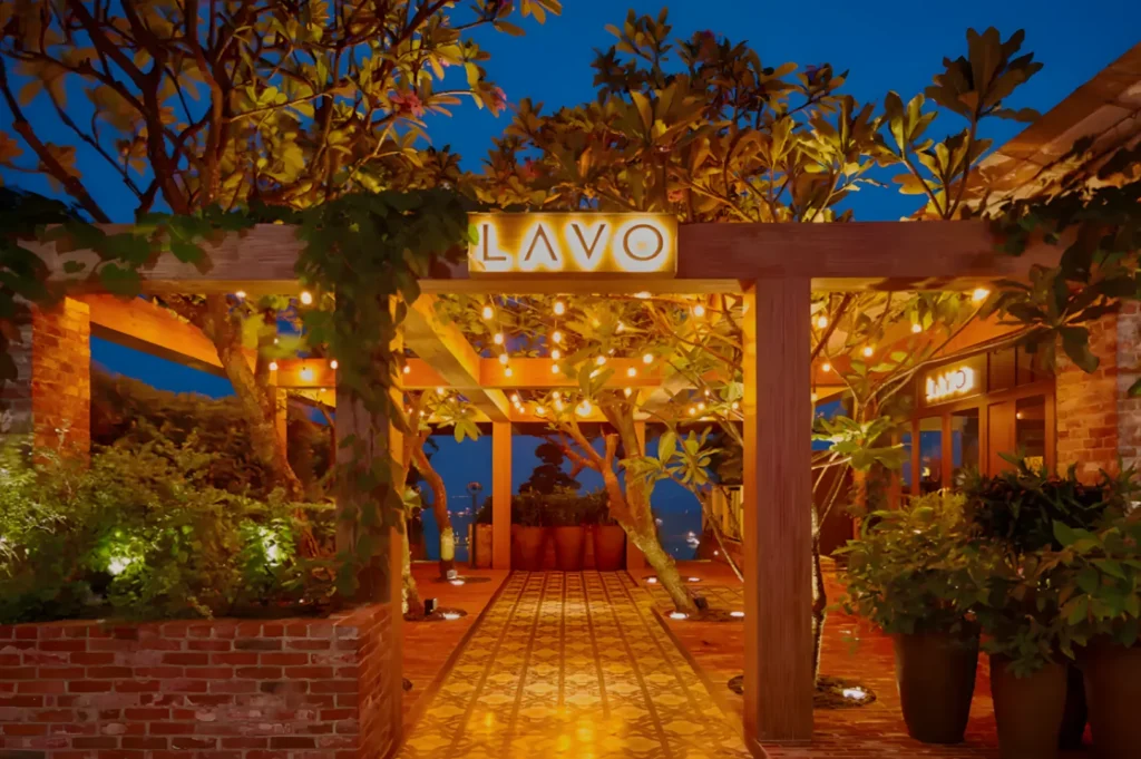 A serene restaurant entrance at dusk, featuring warm string lights, lush foliage, and a patterned tile path, exuding an inviting and cozy ambiance.