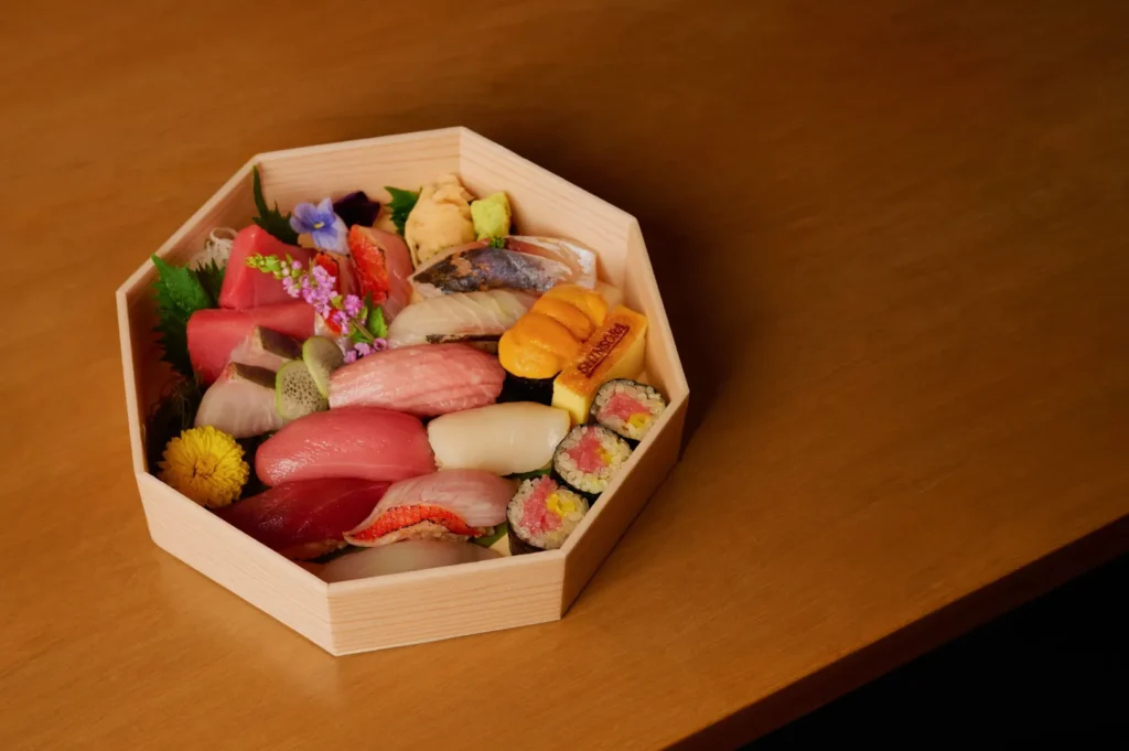 Octagonal wooden box with assorted sushi, including nigiri and rolls, garnished with vibrant flowers and leaves, on a light wooden table.