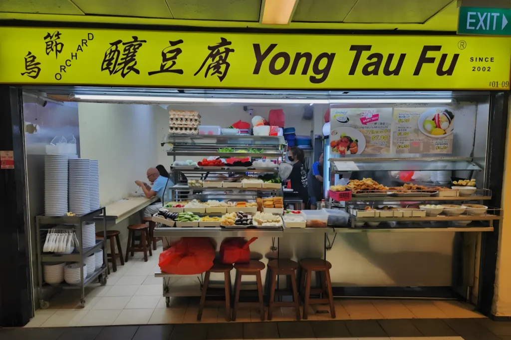 Front view of a colorful Yong Tau Fu food stall with a variety of ingredients displayed. A lone diner sits on a stool, creating a casual, inviting atmosphere.