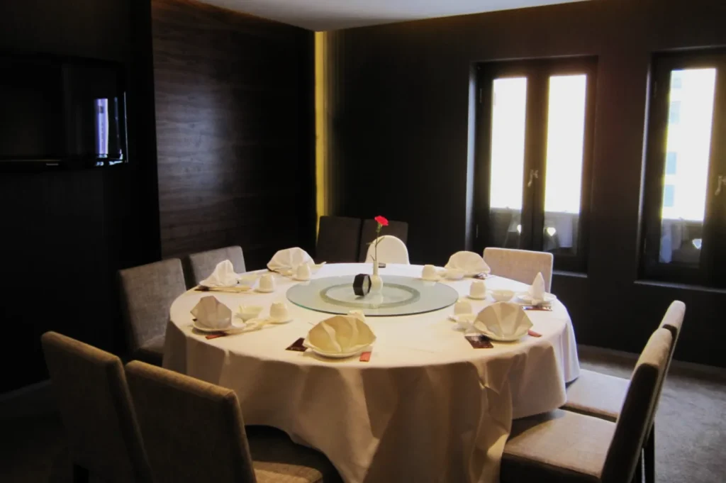 Elegant dining room with a round table set for eight, featuring a white tablecloth, napkins, and a single red flower centerpiece, lit by natural light.