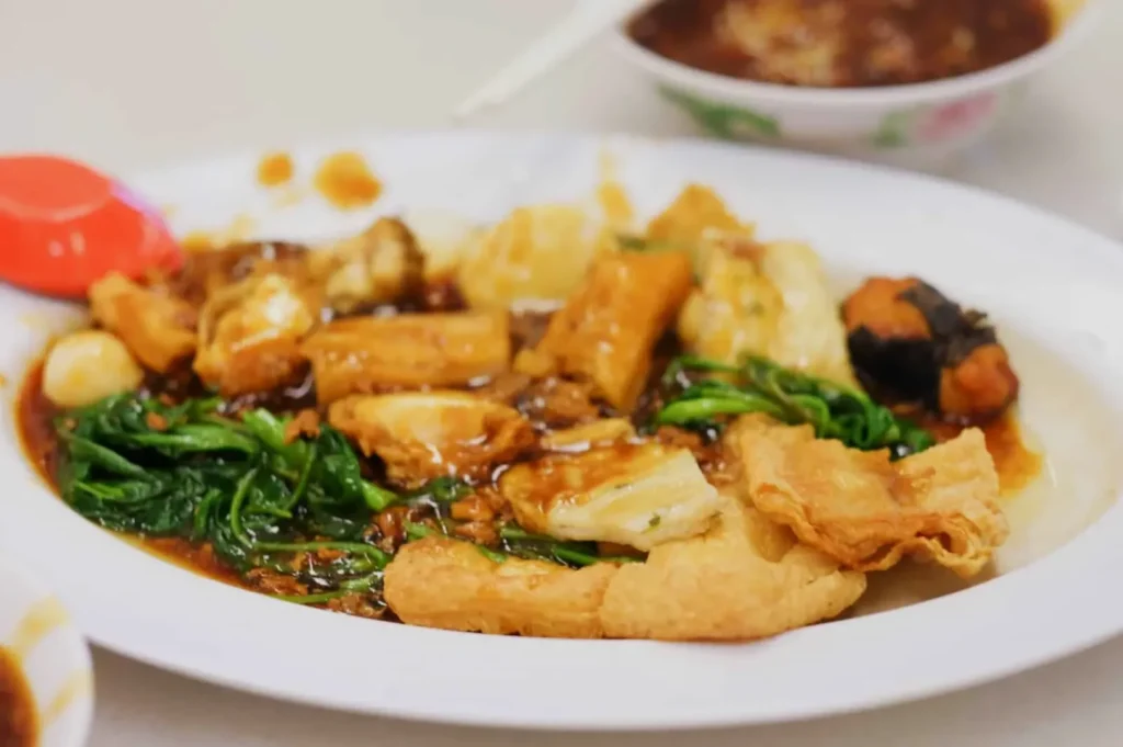 A white plate with yong tau foo, featuring tofu, fish cakes, greens, and sauce. A red spoon rests beside the dish, creating a warm, inviting scene.