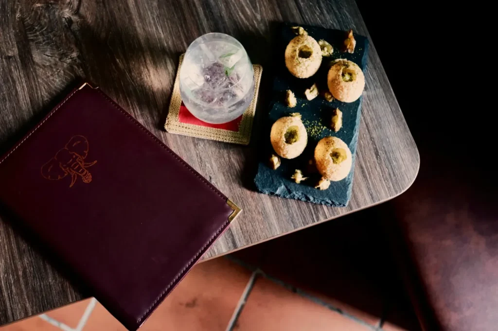 A wooden table with a dark menu, a round cocktail glass on a coaster, and a slate plate with four round appetizers garnished with herbs. Cozy ambiance.
