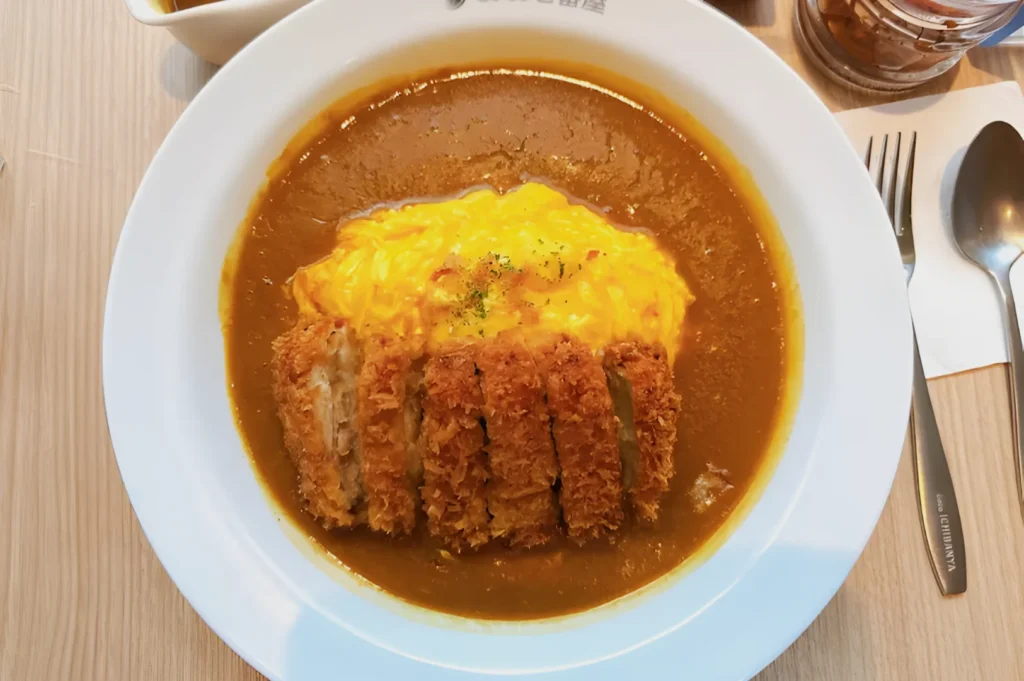A plate of Japanese curry with a golden brown breaded cutlet and creamy egg omelet on top. A spoon and fork are placed on a napkin beside it.