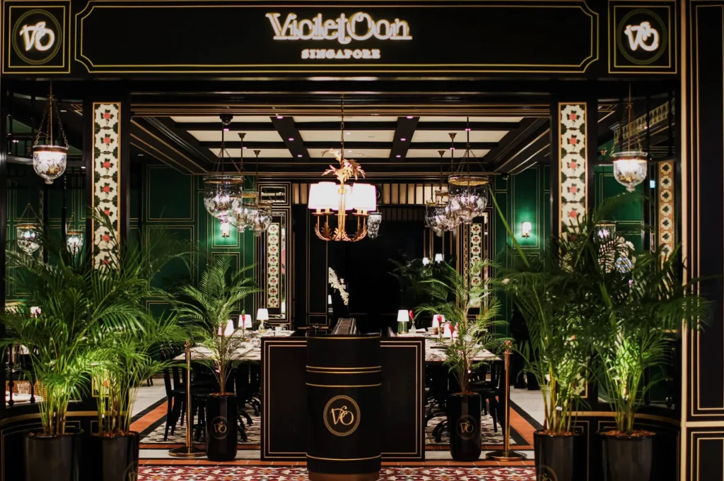 Luxurious restaurant entrance with rich green and gold decor, ornate chandeliers, potted plants, and an elegant, inviting ambiance.