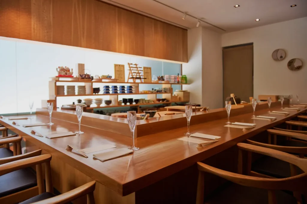 Elegant sushi bar interior with wooden decor, empty counter lined with champagne flutes and chopsticks, creating a serene and inviting atmosphere.