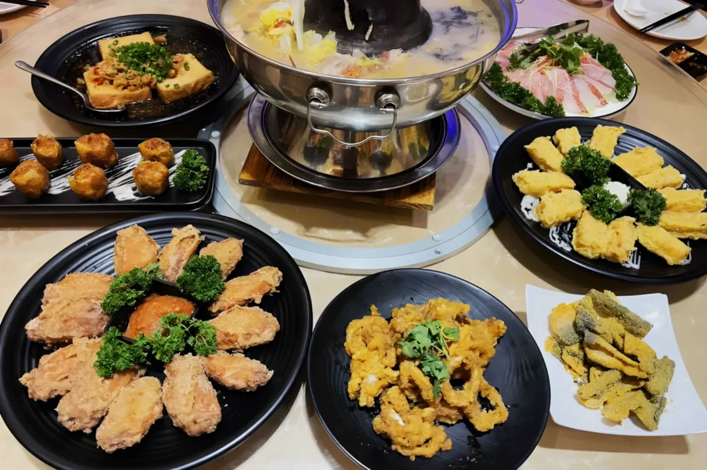A steaming hot pot is centered on a table, surrounded by plates of fried chicken, tofu, tempura, sliced fish, and vegetables, creating a warm, inviting meal.