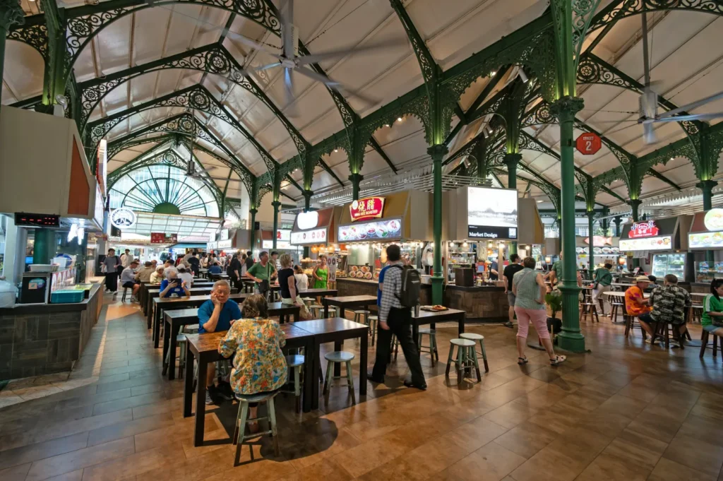 Spacious food court with ornate green iron pillars and high ceilings. People sit at wooden tables surrounded by various brightly lit food stalls.