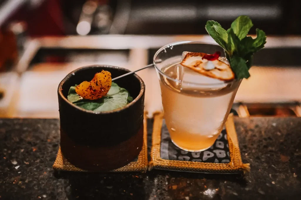 Two artisanal cocktails on a bar counter. Left: a dark round cup with a leaf and orange garnish. Right: clear glass with a dried slice and mint.