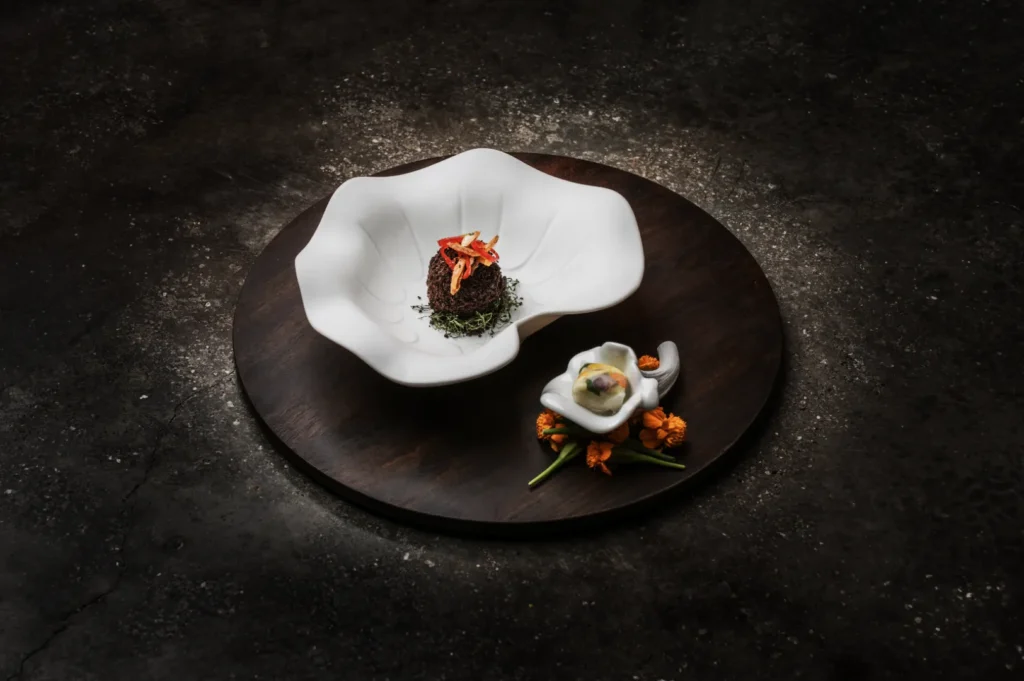 A beautifully plated dish featuring a white shell-like bowl with a small serving of artistic food garnished with herbs and flowers on a wooden platter.