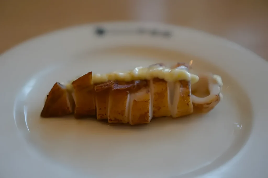 A sliced, grilled squid on a white plate, topped with creamy sauce. The dish is elegantly presented, conveying a fine dining aesthetic.