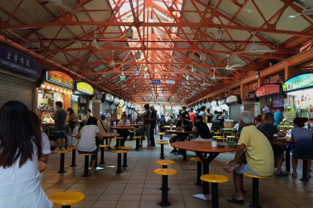 Bustling food court with red beams, filled with people at tables, vibrant food stalls on both sides, and a warm, lively atmosphere.