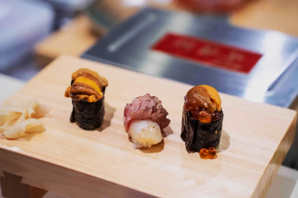 Assorted sushi platter on a wooden board includes two sea urchin gunkan maki and a seared fish nigiri, garnished with pickled ginger on the side.