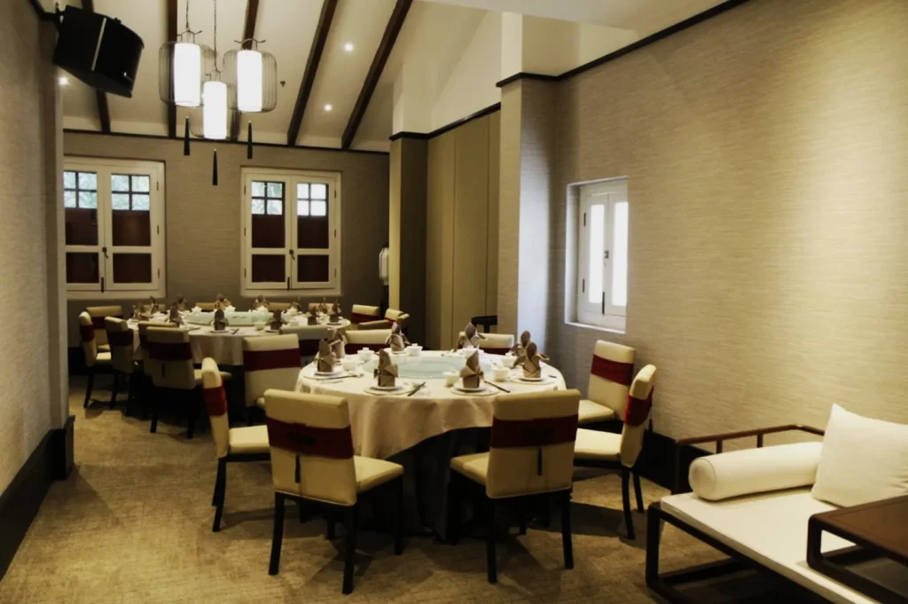 Elegant dining room with round tables set for a meal. Soft lighting, neutral walls, and wooden beams create a cozy, formal ambiance.