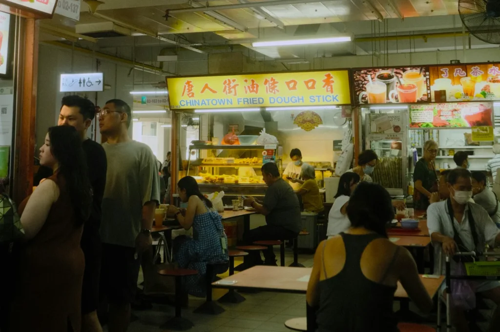 A bustling food stall scene in Chinatown, featuring people ordering and dining beneath a bright sign that reads "Chinatown Fried Dough Stick." The atmosphere is lively.