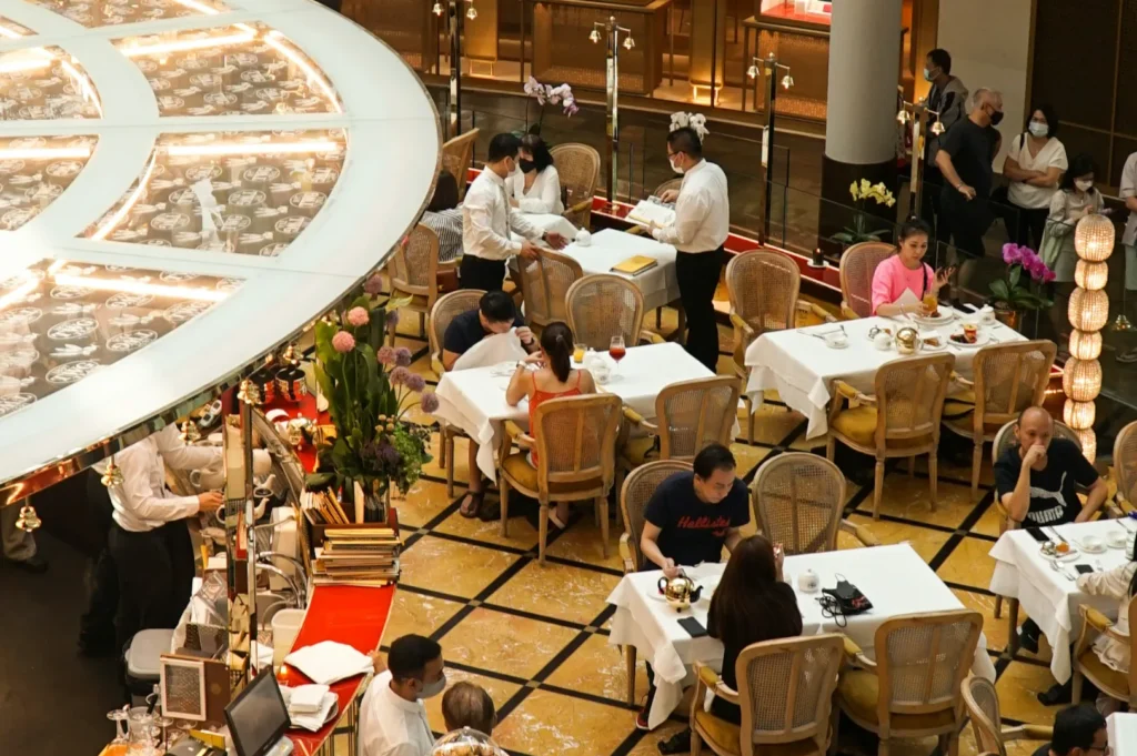 Aerial view of an elegant restaurant with multiple diners at white tablecloth-covered tables. Waitstaff in uniforms attend to guests. The setting feels lively and sophisticated.