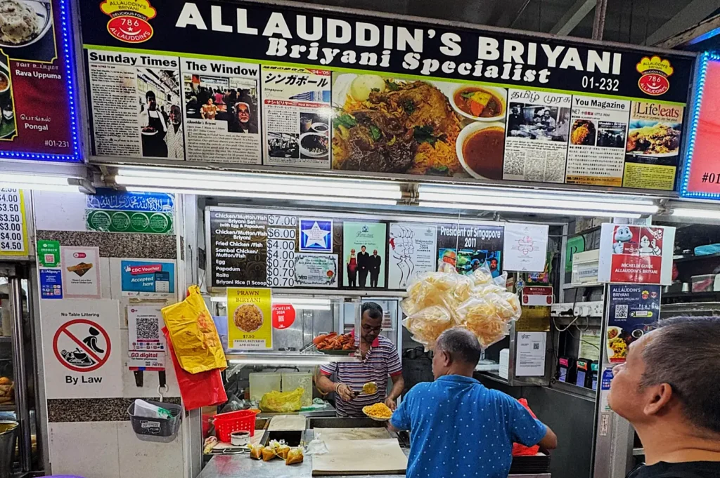 Stall named "Allauddin's Briyani Specialist" with a colorful sign and newspaper clippings. Two men in front, one preparing food. Air of bustling activity.