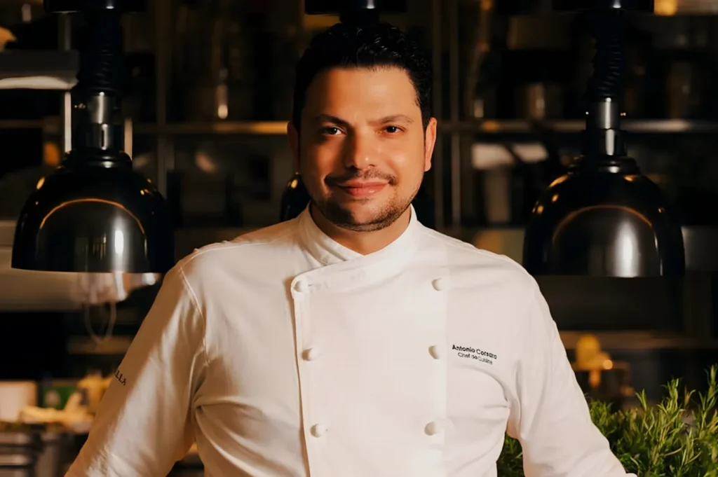 Chef in a white uniform smiles confidently in a warm-lit kitchen, surrounded by hanging lamps and blurred culinary tools, conveying an inviting atmosphere.