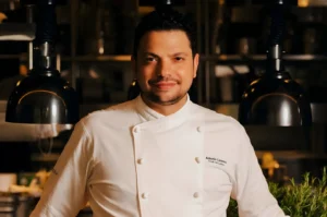 Chef in a white uniform smiles confidently in a warm-lit kitchen, surrounded by hanging lamps and blurred culinary tools, conveying an inviting atmosphere.