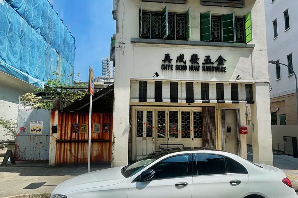 A white, three-story building with "Chye Seng Huat Hardware" signage, open green shutters, a parked white car, and neighboring construction.