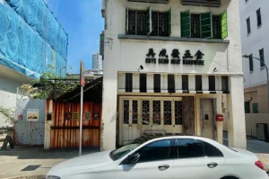 A white, three-story building with "Chye Seng Huat Hardware" signage, open green shutters, a parked white car, and neighboring construction.