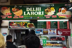 A vibrant food stall sign reads "Delhi Lahori," offering Indian and Pakistani cuisine. Dishes like butter chicken are displayed with prices. Customers stand in line, creating a lively atmosphere. Social media icons suggest online presence, while Michelin Guide mention implies recognition.