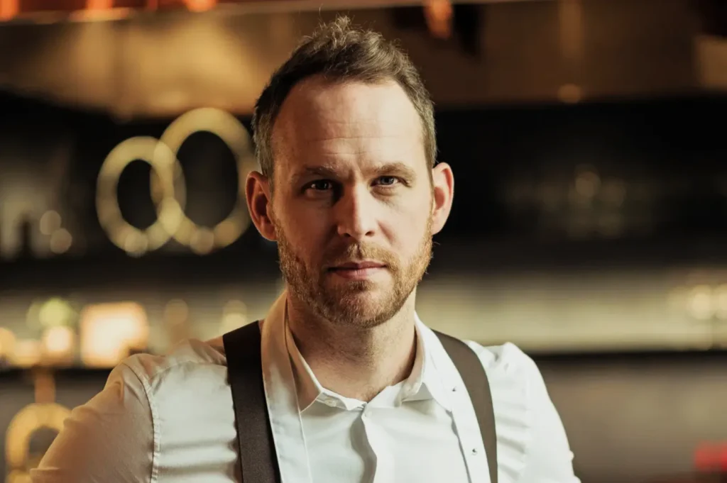 A man in a white shirt with suspenders stands in a dimly lit kitchen, appearing focused and confident. Blurred circles of light add a warm ambiance.