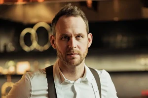 A man in a white shirt with suspenders stands in a dimly lit kitchen, appearing focused and confident. Blurred circles of light add a warm ambiance.